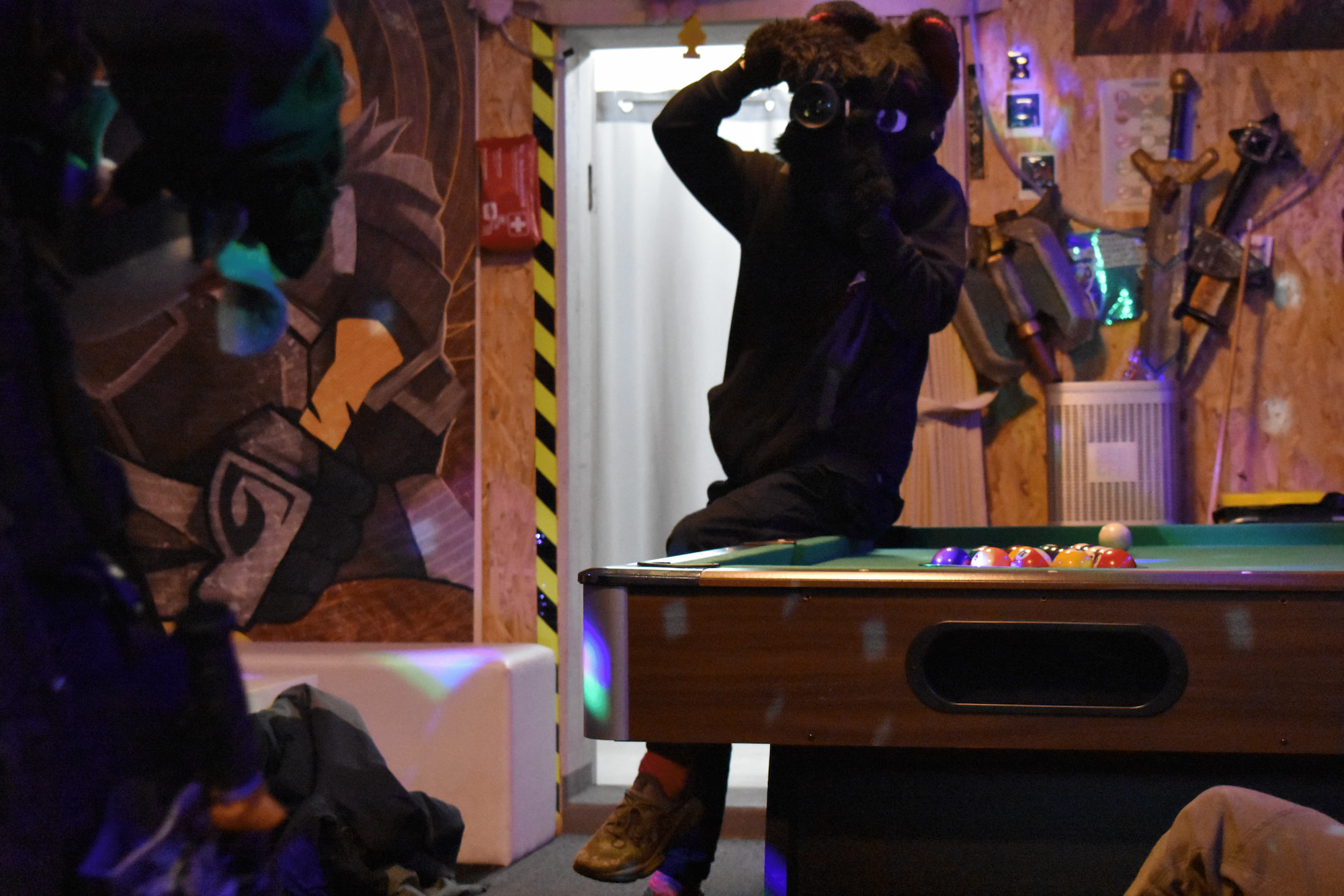 A person in a cat fursuit with purple spots and purple nose with a camera, leaning on a pool table, aiming towards the viewer